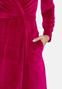 Abito in velluto fucsia con design avvolgente, maniche lunghe, cintura da legare e tasche laterali. Texture liscia con una lucentezza lussuosa.