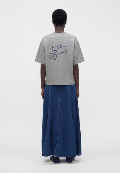 Personne debout de dos, portant un t-shirt gris à manches courtes avec un texte bleu et une longue jupe en jean bleu, sur un fond clair uni.