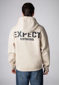 Cremefarbener Hoodie mit großem schwarzem "EXPECT"- und "NOTHING"-Schriftzug auf der Rückseite. Verfügt über eine Kapuze mit Kordelzug, Ärmelbündchen und einen gerippten Saum.