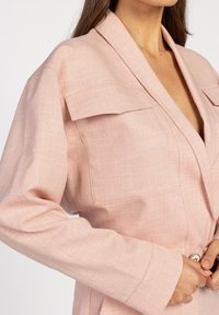 Lichtroze blazer van een gestructureerde stof, diepe V-hals, grote zakken en een knoopsluiting. Heeft gestructureerde schouders en lange mouwen.