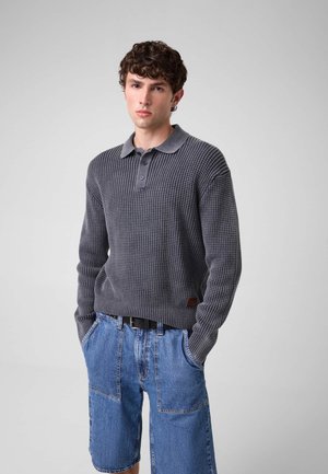 Junger Mann mit lockigem Haar, der ein graues, strukturiertes Langarm-Poloshirt und blaue Denim-Shorts trägt, mit den Händen in den Taschen vor einem schlichten Hintergrund.
