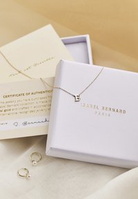 Gold necklace with a letter "B" pendant, displayed on a cream fabric. Includes a certificate of authenticity and pearl-accented earrings in a box.