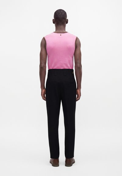 Homme debout de dos, portant un haut rose sans manches, un pantalon noir taille haute et des chaussures brunes sur un fond uni.