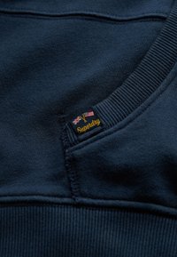 Marineblauwe sweatshirt met een geribde kraag en mouwen, voorzien van een geborduurd "Superdry" label en kleine details van de Britse en Amerikaanse vlag. Zachte textuur.