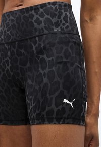 Κοντινό πλάνο ενός ατόμου που φοράει αθλητικές σορτς μαύρου και σκουρόγκρι λεοπάρ μοτίβο με λευκό λογότυπο Puma στο αριστερό μηρό.