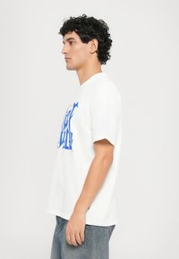 White t-shirt with short sleeves featuring blue graphic text design. Smooth cotton fabric, relaxed fit, rounded neckline, and minimalistic style.