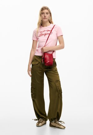Young woman wearing a pink logo t-shirt, olive cargo pants, beige sneakers, and a small red crossbody bag, standing against a white background.