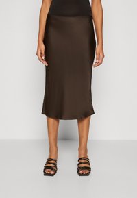 Jupe midi en satin marron avec une texture lisse, un ourlet légèrement évasé et une taille ajustée. Portée avec des sandales à talons hauts noires et à lanières.