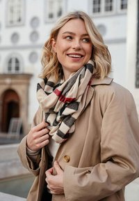 Beige huivi, jossa on musta ja punainen ruutukuosi, roikkuvana kaulassa. Yhdistettynä vaaleaan takkiin, joka on valmistettu pehmeästä, tekstuurisesta kankaasta.