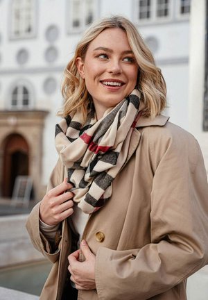 Beige huivi, jossa on musta ja punainen ruutukuosi, roikkuvana kaulassa. Yhdistettynä vaaleaan takkiin, joka on valmistettu pehmeästä, tekstuurisesta kankaasta.