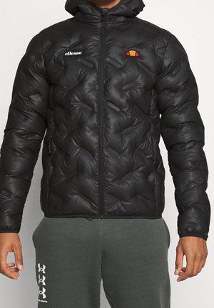 Mężczyzna noszący czarną pikowaną kurtkę z kapturem z logo ellesse oraz ciemnoszare spodnie dresowe z logo Under Armour na lewej nodze.