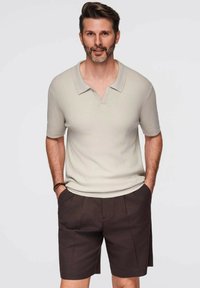 Světle béžové žebrované pólo tričko s límečkem, spárované s tmavě hnědými tailored šortkami. Model stojí s rukama v kapsách a ukazuje správný střih oblečení.