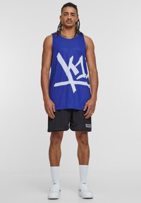 Canotta blu in rete con design grafico bianco, abbinata a pantaloni corti neri con testo bianco. Modello che indossa sneakers bianche e calzini corti.