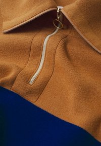 Pull en tricot marron avec une section inférieure bleue, doté d'une fermeture éclair à demi et d'un accent en forme d'anneau doré. Texture douce.