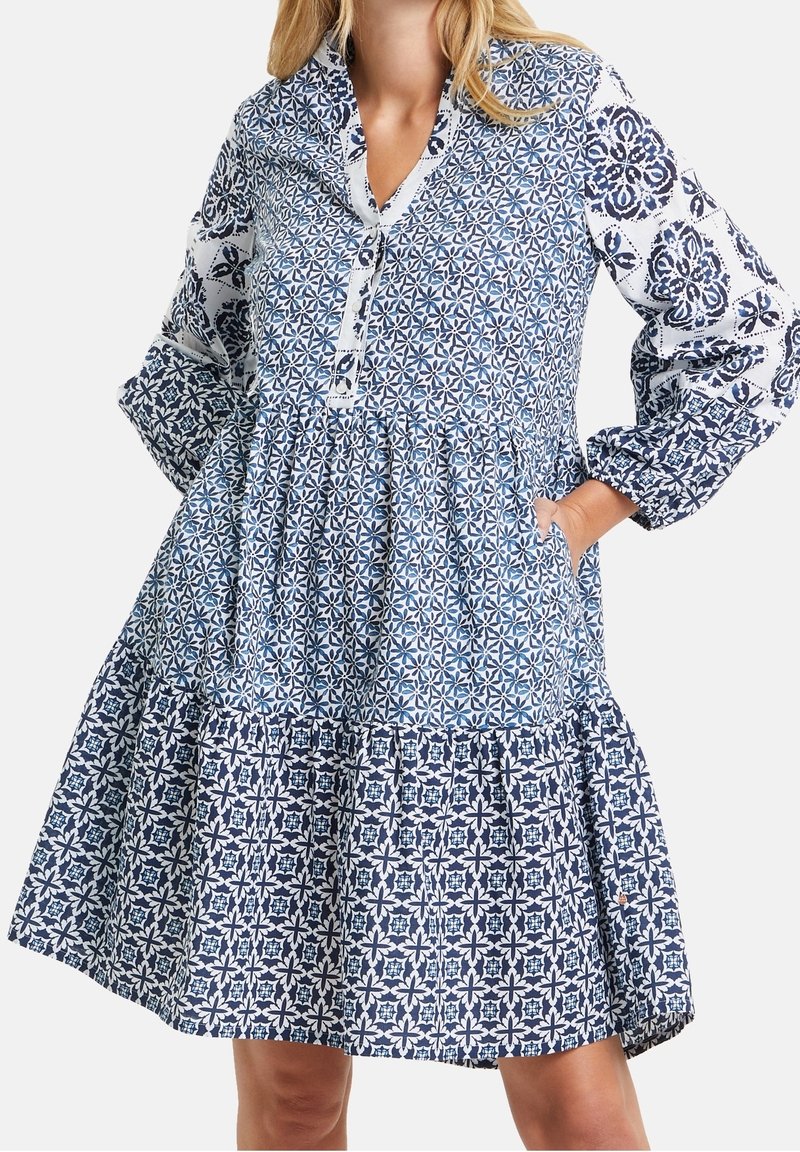 Femme portant une robe mi-longue avec des motifs géométriques floraux bleus et blancs mélangés et des manches longues bouffantes.