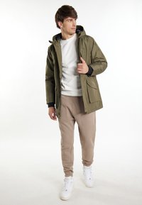 Giacca impermeabile verde oliva con cappuccio, zip frontale e tasche laterali, indossata sopra un maglione bianco e pantaloni jogger beige, abbinata a sneakers bianche.