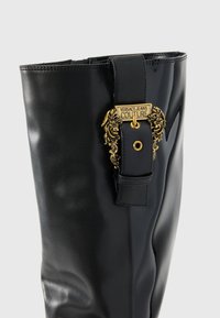 Zwarte glanzende laars tot de knie met een decoratieve gouden gesp en strap met gravure. Gladde textuur met een slanke silhoeet.
