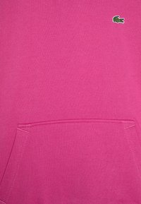 Sudadera rosa con bolsillo frontal, textura suave y un pequeño logo bordado verde en la parte superior izquierda.