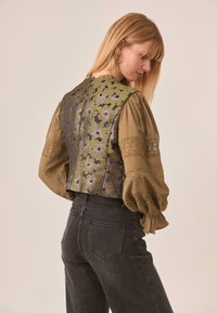 Blusa verde caqui con mangas abullonadas, presenta patrones florales en púrpura y un intrincado detalle de encaje en las mangas. Se muestra la vista trasera.