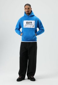 Sweat à capuche bleu avec un graphique d'étiquette blanche, associé à un pantalon noir ample et des chaussures noires épaisses avec des lacets colorés.