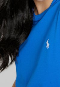 Jasně modré bavlněné tričko s kulatým výstřihem a malým bílým vyšívaným logem na levé straně hrudi. Hladká textura, krátké rukávy.