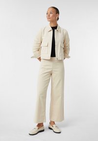 Lichtbeige ritsjack met twee voorzakken, gecombineerd met bijpassende wijde broek. Zwarte top eronder en lichte schoenen met een lage hak.