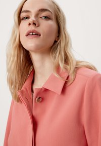 Koraalroze jas met een kraag, sluiting met één knop en een gladde textuur. Gelaagd materiaal met subtiele stikdetails.