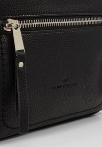 Sac en cuir noir texturé featuring une fermeture éclair en métal argenté, logo embossé sur le devant, et un petit tirant en cuir.