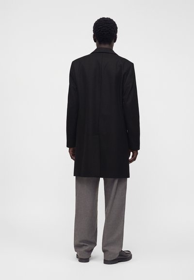 Homme debout, de dos, portant un long manteau noir, un pantalon gris et des chaussures noires sur un fond blanc uni.