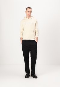 Sudadera con capucha beige, con bolsillo frontal y logo en relieve, combinada con pantalones negros ajustados y zapatos negros con cordones.