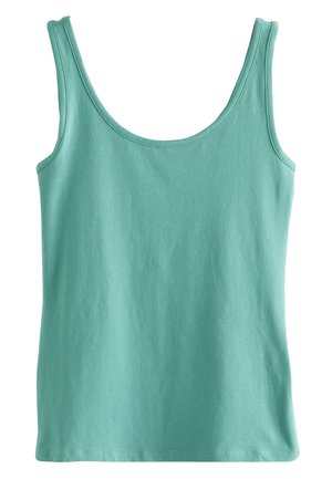 Mujer con cabello rubio largo, que lleva una camiseta sin mangas de color verde azulado, de pie con los brazos cruzados, mirando hacia un lado bajo una luz suave del sol.