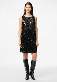 Crna haljina bez rukava prekrivena okruglim, sjajnim šljokicama. A-linijski kroj s valovitim rubom. Model nosi crne kožne čizme do koljena.