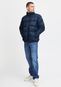 Marineblå dunjakke med høj krave, horisontalt quiltet design, lynlås lukning, sidelommer, parret med blå jeans og hvide sneakers.