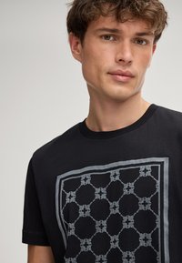 Zwarte katoenen t-shirt met een rechthoekig, grijs geometrisch patroon op de voorkant. Heeft een ronde hals en korte mouwen.