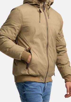 Homme portant une veste à capuche beige avec des poches zippées et des manches retroussées sur un jean bleu, sur un fond uni.