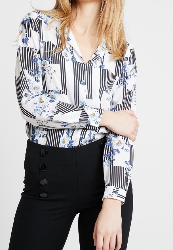Blusa floreale con strisce verticali bianche e nere, maniche lunghe e colletto. Abbinata a pantaloni neri a vita alta con bottoni.