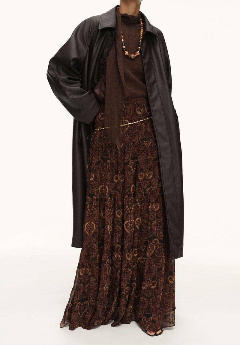 Femme portant un long manteau en cuir marron foncé, une jupe longue marron à motifs, un haut marron, un collier de perles et une fine ceinture chaîne dorée.