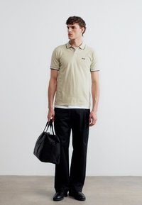 Jeune homme portant un polo beige et un pantalon noir, tenant un sac de sport noir, se tenant contre un mur blanc uni.
