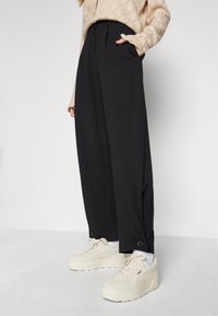 Black wide-leg trousers with front pleats, side pockets, and button accents; paired with beige platform sneakers.