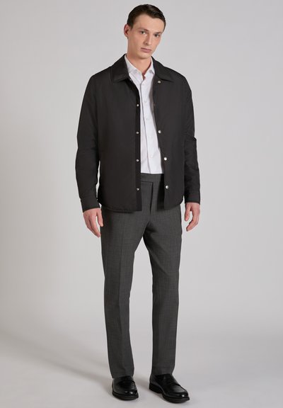 Młody mężczyzna w czarnej zapinanej na guziki kurtce, białej koszuli, szarych spodniach w kratę oraz czarnych skórzanych butach, stojący na jednolitym tle.