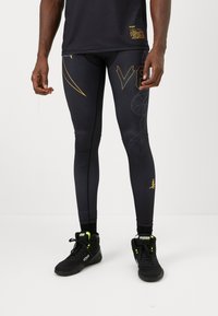 Schwarze Sportleggings mit goldenen Akzenten und Mustern, die ein enges Design aufweisen. Kombiniert mit schwarzen Sneakers mit neongrünen Details.