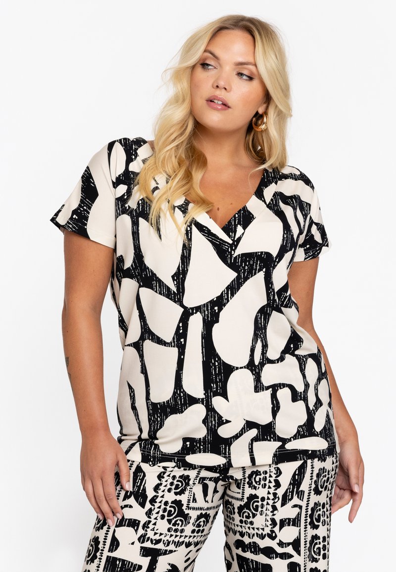 Kurzärmliges Oberteil in Creme mit auffälligem schwarzem abstrakten Muster, V-Ausschnitt und weicher Textur. Kombiniert mit passenden gemusterten Hosen.