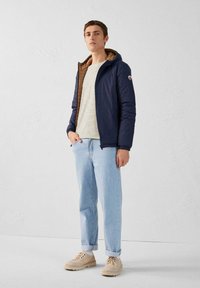 Marine pufferjack met een bruine binnenkant, gestreept shirt, lichtblauwe jeans en beige sneakers. Voorzien van een capuchon en zijzakken.