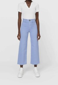 Jeans de pernera ancha en azul claro con cintura alta, que cuentan con un cierre de botón y dobladillo desflecado, combinados con una blusa blanca de manga corta y zapatillas blancas.