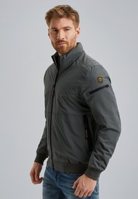 Chaqueta bomber gris con un alto cuello, cierre de cremallera en la parte delantera, bolsillos laterales y un parche de logo en el hombro. Textura suave y lisa, diseño ligero.