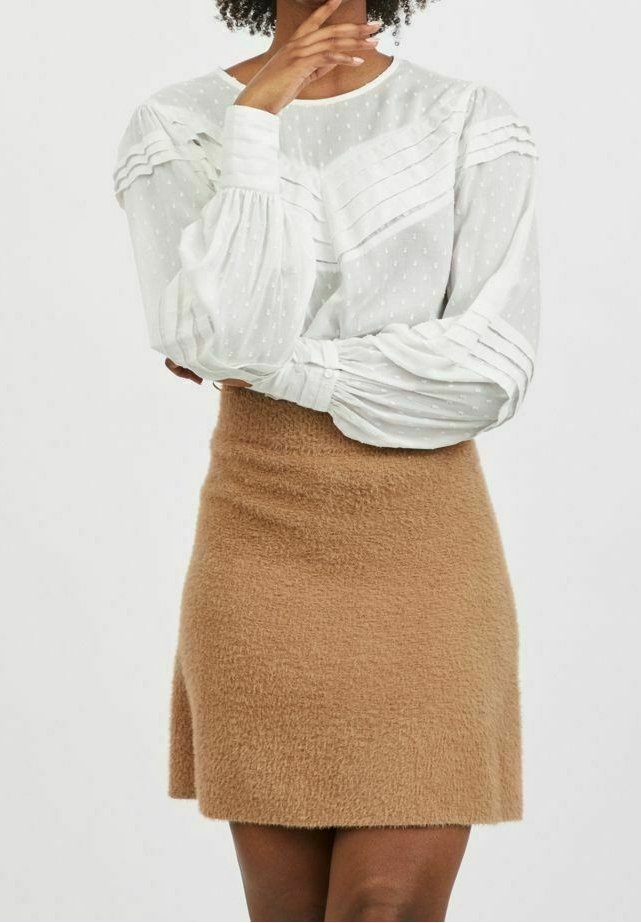 Femme portant un chemisier blanc texturé à manches longues avec des plis et une jupe douce marron longueur genou, posant avec une main près du menton.