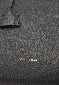 Svart läderväska med en pebblad struktur, prydd med en guld "Coccinelle"-logotyp och en distinkt lädertag som är fäst.