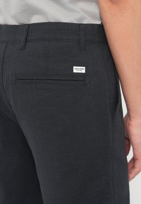 Κοντινό πλάνο ενός ατόμου που φοράει σκούρο ναυτικό μπλε παντελόνι με πίσω τσέπη welt και μια μικρή λευκή ετικέτα της μάρκας Jack & Jones πάνω από την τσέπη.