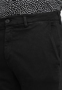 Pantalones de mezclilla negros con un acabado ligeramente texturizado, que presentan un cierre de botón estándar y un diseño clásico de bolsillos.