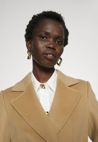 Beige wollen jas met een notchkraag, gecombineerd met een witte shirt met een kleurrijke geometrische stropdas. Gouden spiraal oorbellen zijn zichtbaar.